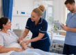 A nurse checks a patient’s identification wristband while another healthcare professional reviews her care notes in a clean hospital ward.