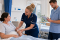 A nurse checks a patient’s identification wristband while another healthcare professional reviews her care notes in a clean hospital ward.